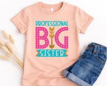 Load image into Gallery viewer, A pink t-shirt with a graphic design that says "PROFESSIONAL BIG SISTER" in pink and teal colors, along with a gold arrow. The t-shirt is placed next to a pair of blue jeans.
