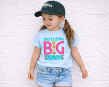 Load image into Gallery viewer, A young girl wearing a blue t-shirt with a graphic design and a black baseball cap stands against a white brick wall.