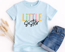 Load image into Gallery viewer, A light blue t-shirt with the text "LITTLE Sister" printed on it, along with a pair of denim shorts.