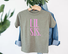 Load image into Gallery viewer, A gray t-shirt with the text "LIL SIS" printed in pink on it, hanging on a wooden hanger.