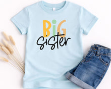 Load image into Gallery viewer, A light blue t-shirt with the text "BIG SISTER" printed on it, along with a pair of denim shorts.
