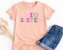 Load image into Gallery viewer, A peach-colored t-shirt with the text "BIG SISTER" printed on it, along with a pair of blue denim shorts, are laid out on a white background.