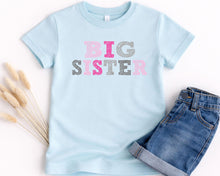 Load image into Gallery viewer, A light blue t-shirt with the text "BIG SISTER" printed on it, along with a pair of denim shorts.