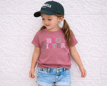 Load image into Gallery viewer, A young girl wearing a green baseball cap and a pink t-shirt with the text "BIG SISTER" is leaning against a white brick wall.
