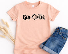 Load image into Gallery viewer, A peach-colored t-shirt with the text "Big Sister" printed on it, along with a pair of blue denim shorts.