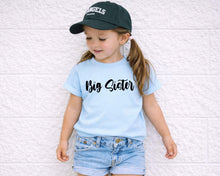 Load image into Gallery viewer, A young girl wearing a blue t-shirt with the text "Big Sister" and a black baseball cap stands against a white brick wall.