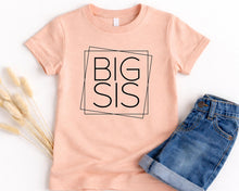 Load image into Gallery viewer, A peach-colored t-shirt with the text "BIG SIS" printed on it, along with a pair of blue denim shorts.