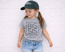Load image into Gallery viewer, A young girl wearing a green baseball cap and a gray t-shirt with the text "BIG SIS" stands against a white brick wall.