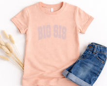 Load image into Gallery viewer, A peach-colored t-shirt with the text "BIG SIS" printed on it, along with a pair of blue denim shorts, are laid out on a white background.
