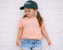Load image into Gallery viewer, A young girl wearing a green baseball cap, a pink t-shirt with the text "BIG SIS", and denim shorts is leaning against a white brick wall.
