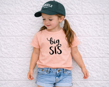 Load image into Gallery viewer, A young girl wearing a green baseball cap, a pink t-shirt with the text "big SIS", and denim shorts is standing against a white brick wall.