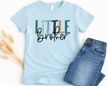 Load image into Gallery viewer, A light blue t-shirt with the text "Little brother" printed on it, along with some jeans and dried flowers.