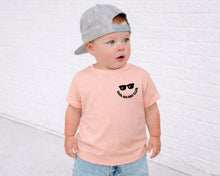 Load image into Gallery viewer, A young child wearing a gray cap and a pink t-shirt with a graphic design stands in front of a white brick wall.