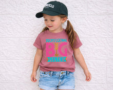 Load image into Gallery viewer, A young girl wearing a green baseball cap and a pink t-shirt with a graphic design stands against a white brick wall.