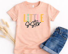 Load image into Gallery viewer, A pink t-shirt with the text "LITTLE SISTER" printed on it, along with a pair of blue jeans.