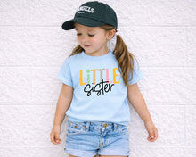 Load image into Gallery viewer, A young girl wearing a blue t-shirt with the text "Little Sister" and a black baseball cap stands against a white brick wall.