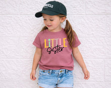 Load image into Gallery viewer, A young girl wearing a pink t-shirt with the text "Little Sister" and a green baseball cap stands against a white brick wall.