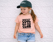Load image into Gallery viewer, A young girl wearing a green baseball cap, a pink t-shirt with the text "COOL BIG SIS CLUB", and denim shorts is leaning against a white brick wall.