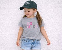 Load image into Gallery viewer, A young girl wearing a green baseball cap and a gray t-shirt with the text "BIG SISTER" stands against a white brick wall.