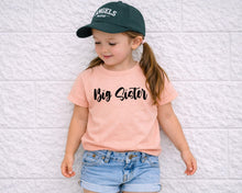 Load image into Gallery viewer, A young girl wearing a green baseball cap, a pink t-shirt with the text "Big Sister", and denim shorts is standing against a white brick wall.