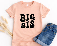 Load image into Gallery viewer, A pink t-shirt with the text "BIG SIS" printed on it, along with a pair of blue jeans, are laid out on a white background.