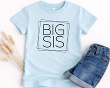 Load image into Gallery viewer, A light blue t-shirt with the text "BIG SIS" printed on it, along with a pair of denim shorts, are displayed on a white background.