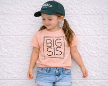 Load image into Gallery viewer, A young girl wearing a green cap and a pink t-shirt with the text "BIG SIS" stands against a white brick wall.