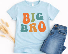 Load image into Gallery viewer, A light blue t-shirt with the text "BIG BRO" printed in orange, green, and orange letters, along with a pair of blue jeans.
