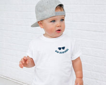 Load image into Gallery viewer, A young child wearing a white t-shirt with a graphic and text, standing in front of a white brick wall.
