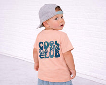 Load image into Gallery viewer, A young child wearing a gray cap and a peach-colored t-shirt with the text "COOL BIG CLUB" printed on it, standing in front of a white brick wall.
