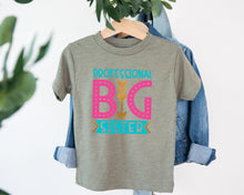Load image into Gallery viewer, A gray t-shirt with a graphic design that says "PROFESSIONAL BIG SISTER" in pink and teal colors, hanging on a wooden hanger.
