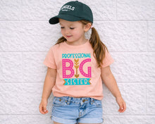 Load image into Gallery viewer, A young girl wearing a green baseball cap, a pink t-shirt with the text "DPROFESSIONAL BIG SISTER" and denim shorts, standing against a white brick wall.
