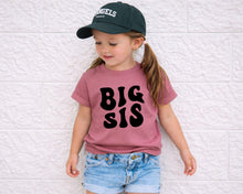 Load image into Gallery viewer, A young girl wearing a green baseball cap and a pink t-shirt with the text "BIG SIS" stands against a white brick wall.
