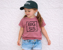 Load image into Gallery viewer, A young girl wearing a green baseball cap and a pink t-shirt with the text "BIG SIS" stands against a white brick wall.
