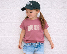 Load image into Gallery viewer, A young girl wearing a green baseball cap and a pink t-shirt with the text "BIG SIS" stands against a white brick wall.
