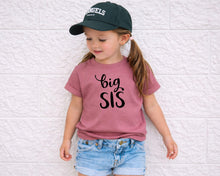 Load image into Gallery viewer, A young girl wearing a green baseball cap and a pink t-shirt with the text "big SIS" stands against a white brick wall.
