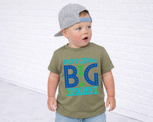 Load image into Gallery viewer, A young child wearing a green t-shirt with a graphic design and a gray baseball cap, standing in front of a white brick wall.
