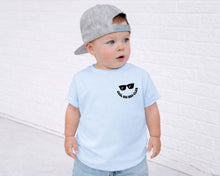 Load image into Gallery viewer, A young child wearing a gray baseball cap and a light blue t-shirt with a smiley face design stands in front of a white brick wall.
