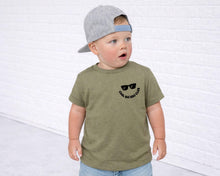 Load image into Gallery viewer, A young child wearing a green t-shirt with a graphic and a gray baseball cap stands in front of a white brick wall.

