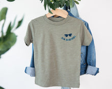 Load image into Gallery viewer, A gray t-shirt with sunglasses printed on it, hanging on a wooden hanger.
