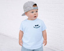 Load image into Gallery viewer, A young child wearing a gray baseball cap and a light blue t-shirt with sunglasses printed on it, standing in front of a white brick wall.
