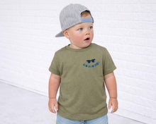 Load image into Gallery viewer, A young child wearing a green t-shirt with a graphic and a gray baseball cap stands in front of a white brick wall.
