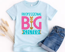Load image into Gallery viewer, A light blue t-shirt with a graphic design that says "PROFESSIONAL BIG SISTER" in pink and teal colors, along with a gold arrow. The t-shirt is placed next to a pair of denim jeans.
