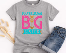 Load image into Gallery viewer, A gray t-shirt with a graphic design that says "PROFESSIONAL BIG SISTER" in pink and teal colors, along with a graphic of a stylized arrow. Next to the t-shirt is a pair of blue jeans.
