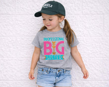 Load image into Gallery viewer, A young girl wearing a green baseball cap and a gray t-shirt with a graphic design stands against a white brick wall.
