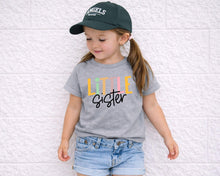 Load image into Gallery viewer, A young girl wearing a green baseball cap and a gray t-shirt with the text "Little Sister" is leaning against a white brick wall.
