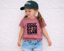 Load image into Gallery viewer, A young girl wearing a green baseball cap and a pink t-shirt with the text "COOL BIG SIS CLUB" stands against a white brick wall.
