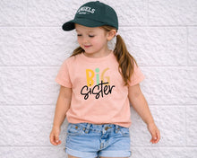 Load image into Gallery viewer, A young girl wearing a green cap and a pink t-shirt with the text "Big Sister" is leaning against a white brick wall.
