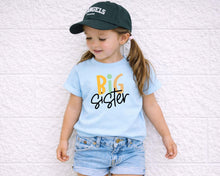 Load image into Gallery viewer, A young girl wearing a blue t-shirt with the text "BIG Sister" and a black baseball cap stands against a white brick wall.
