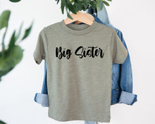 Load image into Gallery viewer, A gray t-shirt with the text "Big Sister" printed on it, hanging on a wooden hanger.
